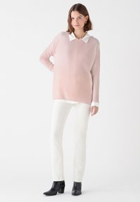Maglione oversize rosa chiaro con colletto bianco, indossato sopra una camicia bianca e abbinato a pantaloni bianchi e stivali neri con tacco.
