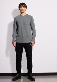 Maglietta grigia a maniche lunghe con taschino sul petto, abbinata a pantaloni neri e stivaletti neri alla caviglia. Tessuto liscio, vestibilità casual.