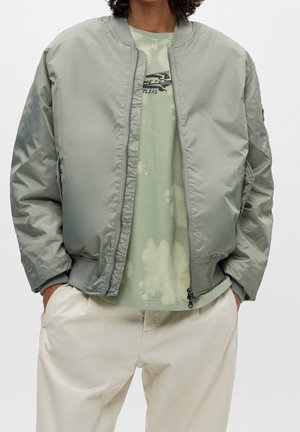 Personne portant une veste bomber gris clair sur un t-shirt graphique tie-dye vert avec un pantalon beige, les mains dans les poches, debout devant un fond blanc.