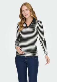Haut à rayures noir et blanc à manches longues avec un col noir uni, présentant un décolleté en V et une texture côtelée, associé à un jean en denim bleu.