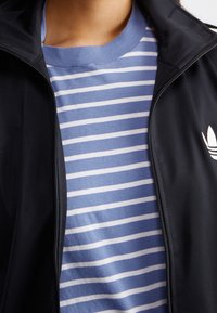 Persona vistiendo una camisa de rayas azul y blanca debajo de una chaqueta negra parcialmente cerrada con una logo blanco de Adidas en el pecho.