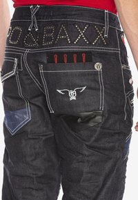 Dunkelblaue Jeans haben dekorative Nähte, mehrere Taschen und metallische Akzente. Auffällige Elemente sind rote Nähte und Logo-Patches.