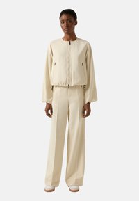 Kremowa kurtka bomberka z zamkiem z przodu, bocznymi kieszeniami i szerokimi rękawami, zestawiona z pasującymi wysokim stanem spodniami. Gładka tekstura.