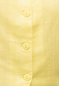 Geltono audinio artimas planas su trimis vienodos spalvos mygtukais, siūtais vertikaliai eilute ant drabužio.