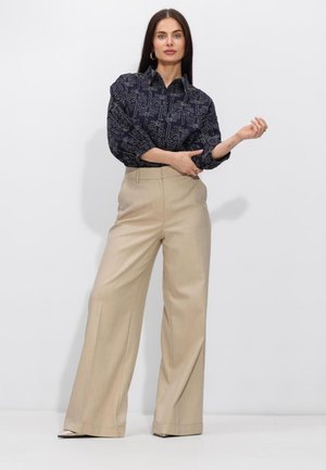 Vrouw met lang donker haar, gekleed in een marineblauw blouse met print en beige, hooggetailleerde wijd uitlopende broek, die haar mouw rechtzet tegen een neutrale achtergrond.