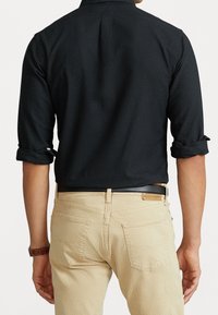 Schwarzes Hemd mit Knopfverschluss und hochgerollten Ärmeln, kombiniert mit hellbeigen Hosen. Der Stoff wirkt glatt, hat ein ordentliches Design und sitzt eng.