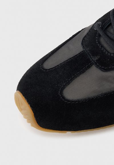 Zapatilla deportiva de ante negro con paneles de malla, puntera redondeada y suela de goma color chicle. Cuenta con cordones y un talón reforzado para mayor soporte.