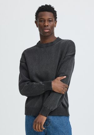 Jeune homme aux cheveux courts et bouclés, portant un pull gris foncé et un jean bleu, debout les bras croisés sur la poitrine.