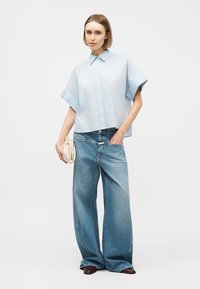 Γυναίκα φορώντας ανοιχτό μπλε πουκάμισο με κοντά μανίκια, φαρδιά μπλε τζιν, κρατώντας μια μπεζ τσάντα φάκελο, στέκεται μπροστά σε απλό φόντο.