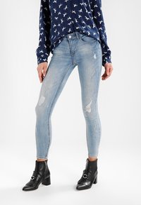 Světle modré skinny džíny se zařezanými nášivkami, kombinované s tmavou vzorovanou blůzou a černými botami na podpatku. Hladká denimo textura je viditelná.