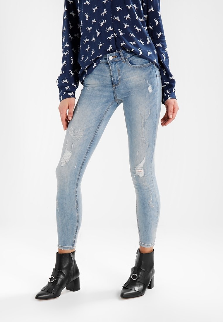 Světle modré skinny džíny se zařezanými nášivkami, kombinované s tmavou vzorovanou blůzou a černými botami na podpatku. Hladká denimo textura je viditelná.