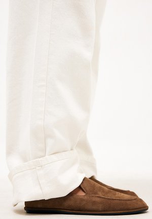 Hnedé semišové slip-on topánky nosené s bielymi nohavicami vyhrnutými na členkoch, zobrazené z boku na úrovni členka.