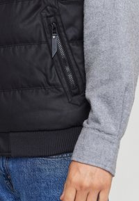Gilet imbottito nero con tasca con zip e manica di maglione grigia. Presenta orlo e polsini a coste, con jeans in denim visibili sotto.