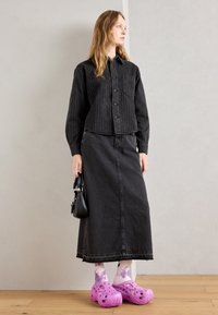Chemise courte noire à rayures, jupe en denim noir, sabots perforés violets et chaussettes tie-dye. Petit sac à main noir à la main.