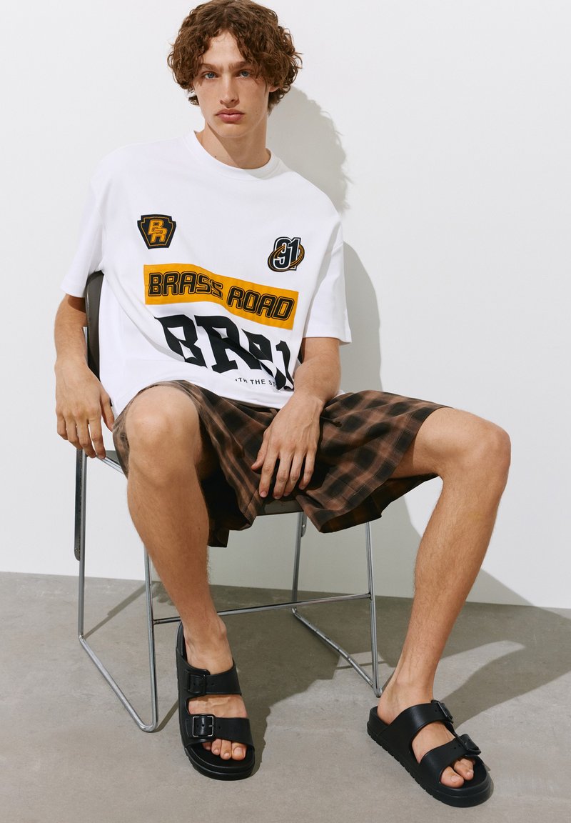 Jeune homme aux cheveux bouclés assis sur une chaise en métal portant un t-shirt blanc à motif graphique, un short à carreaux marron et des sandales noires à boucle.