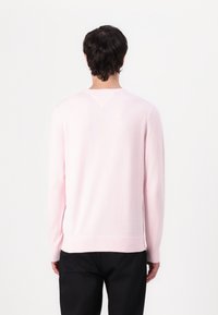 Homme aux cheveux foncés portant un pull rose clair à manches longues et un pantalon noir, debout face à un fond blanc uni.