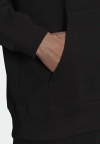 Felpa nera con polsini e fondo a costine, dotata di tasca frontale tipo kangaroo. Il materiale sembra morbido e con una texture.