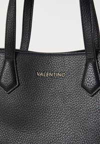 Must musta kott, millel on tekstureeritud pind, kahekordne vastupidav käepide ja kuldne "VALENTINO" logo esiküljel.