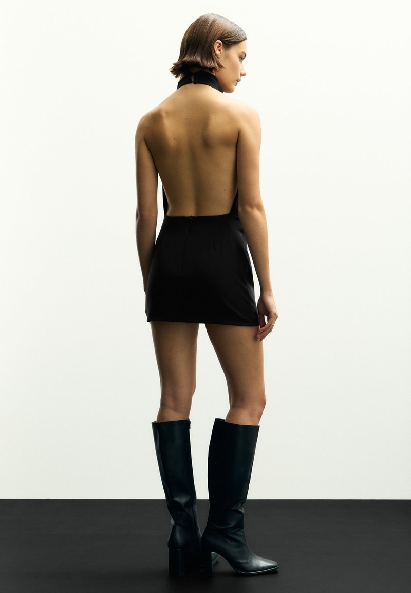 Schwarzes ärmelloses Minikleid mit einem offenen Rücken, kombiniert mit knielangen schwarzen Lederstiefeln mit einem verjüngten Absatz. Glatte Textur.
