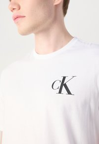 Bijela pamučna majica s okruglim izrezom, s crnim "cK" logotipom na lijevoj strani prsnog područja. Glatka tekstura, jednostavan dizajn.
