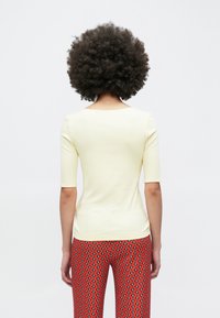 Svetlo rumena prilegajoča majica s tremi četrtinami rokavov, gladke teksture in izrezom v obliki zajemalke, kombinirana z živahnimi, vzorčastimi rdečimi hlačami.