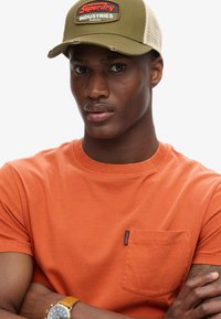 Orange bomulds-t-shirt med en lille lomme og et rødt mærke. Olivengrøn kasket med netbag og et frontmærke. Gul ur på håndleddet.