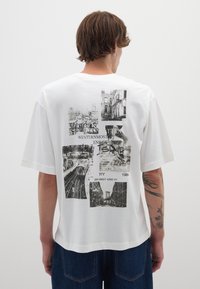 Camiseta de algodón blanca con diseño oversize, que presenta gráficos en negro de monumentos de Nueva York y texto en la parte trasera. Manga corta, corte relajado.