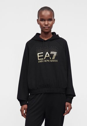 Personne portant un sweat à capuche noir EA7 Emporio Armani avec logo doré et cordons, assorti d’un pantalon noir coordonné, sur un fond uni.