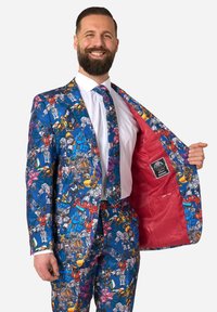 Man smiling, wearing a vibrant blue suit and tie patterned with colorful Transformers characters, showing the red jacket lining with a Transformers patch.