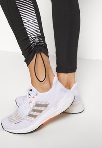 Svarta sportleggings med reflexstripes och justerbara anklar, tillsammans med vita löparskor med grå accenter och texturerade sulor.