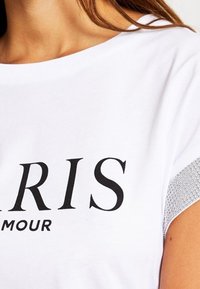 Bílý tričko s černým textem "PARIS" a "GLAMOUR" pod ním. Stříbrné ozdoby na rameni. Krátké rukávy a uvolněný střih.