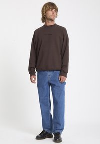 Brązowy sweatshirt z czarnym logo "VOLCOM", w połączeniu z niebieskimi szerokimi dżinsami. Stylizowany z czarnymi sznurowanymi butami, na białym tle.