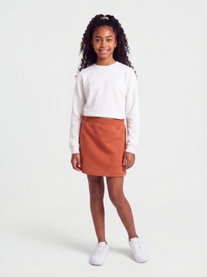 Fille souriante aux longs cheveux bouclés, vêtue d'une chemise blanche à manches longues, d'une jupe orange et de baskets blanches, debout sur un fond uni.