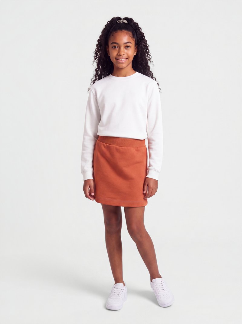 Fille souriante aux longs cheveux bouclés, vêtue d'une chemise blanche à manches longues, d'une jupe orange et de baskets blanches, debout sur un fond uni.