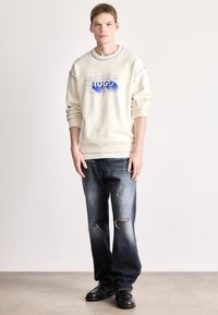 Krém színű pulóver kék "HUGO" grafikus logóval, sötét farmernadrággal, amely kopott részletekkel rendelkezik, és fekete lábbelivel párosítva.