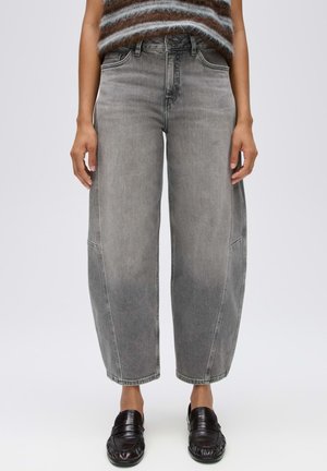 Persoon draagt losse grijze gewassen jeans, gestreepte zachte trui en zwarte instap loafers, staand tegen een witte achtergrond.