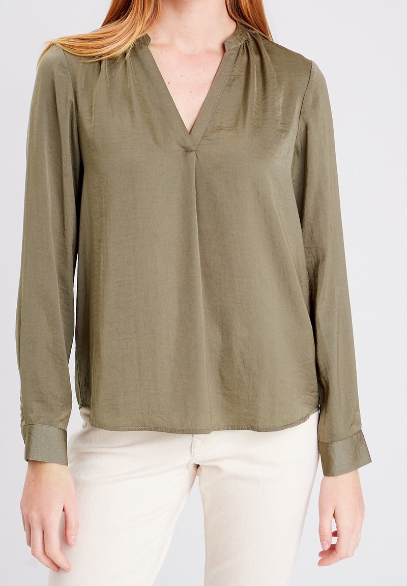 Femme portant un chemisier vert olive à manches longues avec un col en V et un pantalon crème, debout devant un fond clair uni.