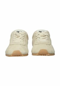 Pari beigeä mokkanahkaisista tennareista, joissa on kumipohjat, nyöritys edessä ja brändätyt kielen merkit, nähtynä edestä.