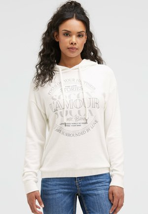 Junge Frau mit lockigem Haar, die einen weißen Hoodie mit Text und blaue Jeans trägt, steht vor einem schlichten, hellen Hintergrund.