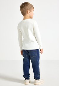 White long-sleeve ribbed shirt paired with dark blue jeans. The jeans feature back pockets and a relaxed fit, worn with light-colored shoes.