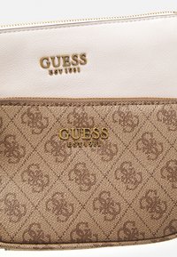 Ruskea kuviollinen käsilaukku, jossa on sileä valkoinen yläosa. Sisältää kultaisen "GUESS"-logon ja vetoketju yksityiskohtia. Teksturoitu pinta ja geometrinen muotoilu.