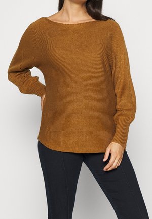 Vrouw draagt een losse, bruingebreide trui met lange mouwen en nauwsluitende zwarte broek, poseert met één hand op haar heup tegen een effen achtergrond.