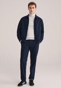 Temno modra zip-up jakna čez belo turtleneck, v kombinaciji s temno modrimi hlačami in temno zelenimi športnimi čevlji z belimi črtami.