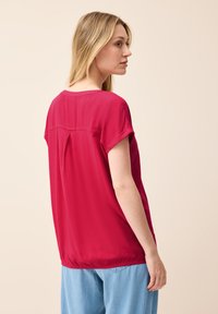 Losen, kurzärmeligen Bluse aus einem hellen pinken Stoff mit glatter Textur. Sie verfügt über eine Rückenfalte und einen runden Ausschnitt und wird mit blauen Jeans kombiniert.