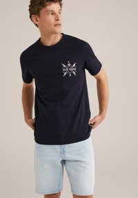 Jūras zila kokvilnas T-kreklu ar grafisku apdruku "BLUE RIDGE" krūtīs, savienots ar gaišziliem džinsu šortiem.