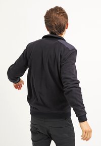 Giovane uomo con capelli castani corti, che indossa una giacca nera e pantaloni neri, rivolto di spalle con le braccia leggermente aperte.
