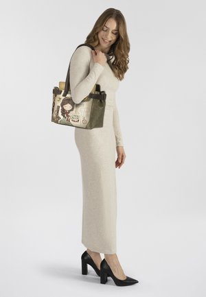 Grüne Lederhandtasche mit Cartoon-Design, zwei Griffen und dekorativen Akzenten. Kombiniert mit einem figurbetonten cremefarbenen Kleid und schwarzen Absätzen.