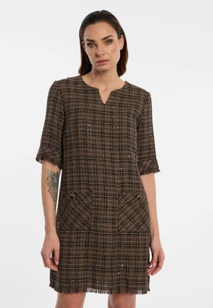 Woman wearing a brown plaid short-sleeve dress with two front pockets and frayed edges, standing against a plain white background.