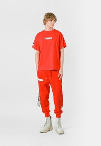 Camiseta de algodón roja con un detalle de bolsillo en el frente, combinada con unos pantalones jogger rojos con cremalleras laterales y acentos de flecos, complementados con botas de combate crema.