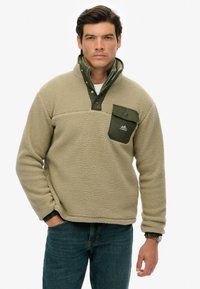Beige fleece pullover med grøn krave og brystlomme, der har en tekstureret overflade og knaplukning. Elastiske manchetter og kant.